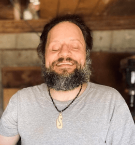 A man with a beard and mustache smiles with his eyes closed. He is wearing a light gray t-shirt and a necklace with a pendant. The background appears to be an indoor space with wooden elements.